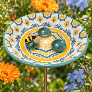 May include: A shallow, round ceramic bird bath dish featuring a teal and yellow geometric pattern. Inside the dish are two smooth, round, teal glass stones and a decorative metal bee figurine with black, yellow, and clear crystal accents.