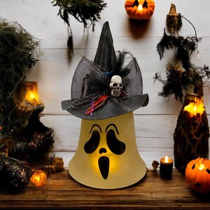 May include: A white ceramic ghost with a black witch hat and a skull decoration. The ghost has a spooky face with two eyes and a mouth. The hat has a black mesh and a black feather. The ghost is lit from within, creating a warm glow.