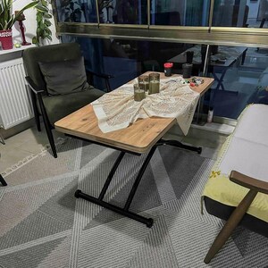 May include: A wooden table with a black metal frame and a white lace tablecloth. The table is set with three candles and a few other items. The table is in a living room setting with a green chair and a rug.