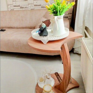 Könnte beinhalten: Ein Beistelltisch aus Holz mit geschwungenem Design. Der Tisch trägt ein weißes Tablett mit einem dekorativen Vogel und eine weiße Kanne mit gelben Tulpen. Ein kleineres Tablett darunter enthält eine kleine Vase und andere dekorative Gegenstände.