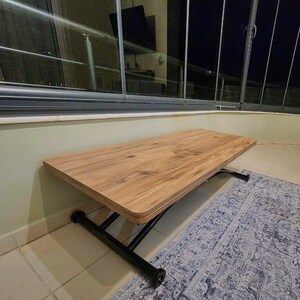 May include: A wooden folding table with a black metal frame. The table is made of light brown wood with a wood grain pattern. The table is folded up and sitting on a rug.