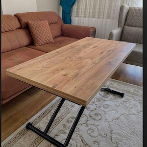 May include: A wooden coffee table with a black metal X-shaped base. The table top is made of light brown wood with a wood grain pattern. The table is sitting on a cream-colored rug with a floral pattern.