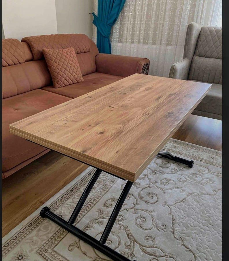 May include: A wooden coffee table with a black metal X-shaped base. The table top is made of light brown wood with a wood grain pattern. The table is sitting on a cream-colored rug with a floral pattern.