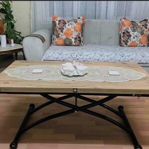 May include: A light brown wooden coffee table with black metal legs that extend to create a larger surface. The table has a white lace tablecloth and a set of white teacups and saucers on a white plate.
