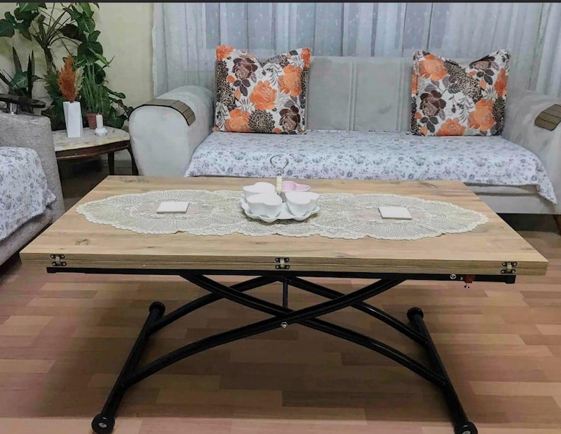 May include: A light brown wooden coffee table with black metal legs that extend to create a larger surface. The table has a white lace tablecloth and a set of white teacups and saucers on a white plate.
