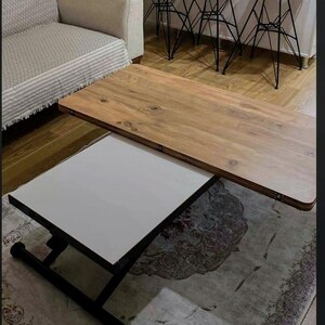 May include: A wooden coffee table with a white top that lifts up to reveal a larger wooden surface. The table has a black metal frame and is sitting on a patterned rug.
