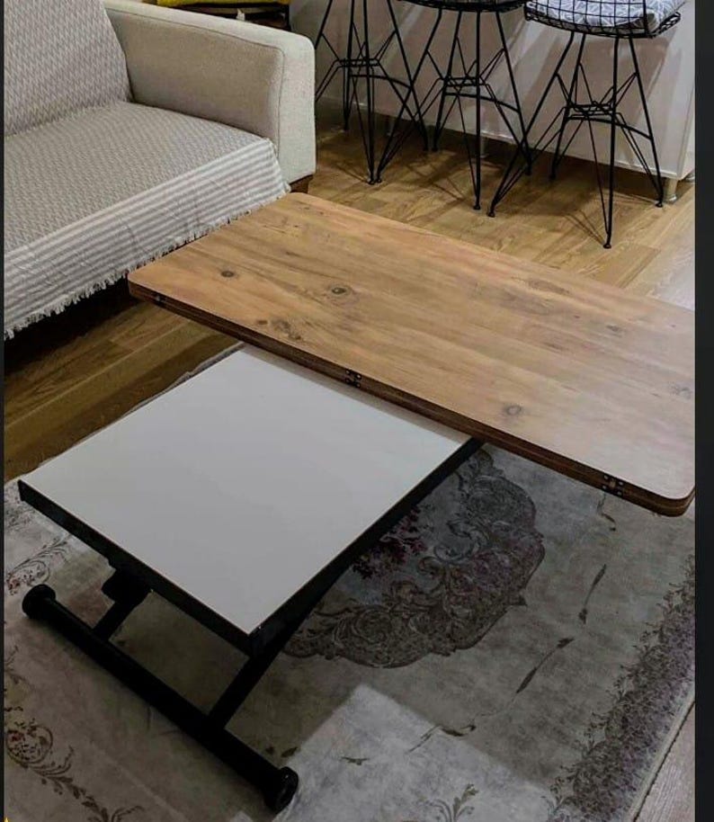 May include: A wooden coffee table with a white top that lifts up to reveal a larger wooden surface. The table has a black metal frame and is sitting on a patterned rug.