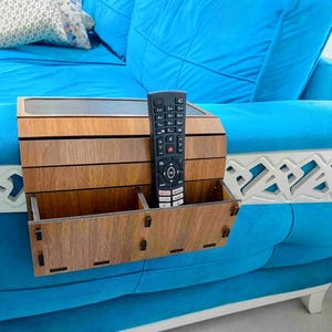 Op de afbeelding: Een houten organizer voor de armleuning van een bank met een ingebouwd compartiment voor een afstandsbediening en twee open opbergvakken. De organizer is gemaakt van lichtbruin hout en is ontworpen om op de armleuning van een bank te worden geplaatst. Een zwarte afstandsbediening bevindt zich in de organizer.