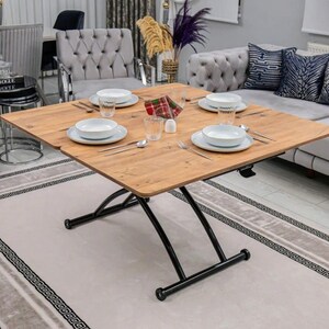 May include: A square wooden table with a black metal base. The table is set with four white plates, four glasses, and a red and black napkin. The table is in a living room setting with a rug and furniture.