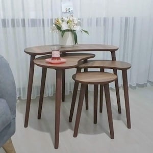 May include: A set of four wooden nesting tables with a dark brown finish. The tables have a unique, organic shape and tapered legs. A vase of white flowers and a glass of tea with a pink saucer are on the tables. The tables are arranged in front of a white curtain.