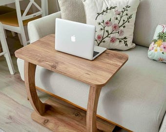 May include: A small, wooden side table with a rectangular top and curved legs. A silver laptop is placed on the table. The table is made of light brown wood and sits next to a cream-colored sofa with decorative pillows.