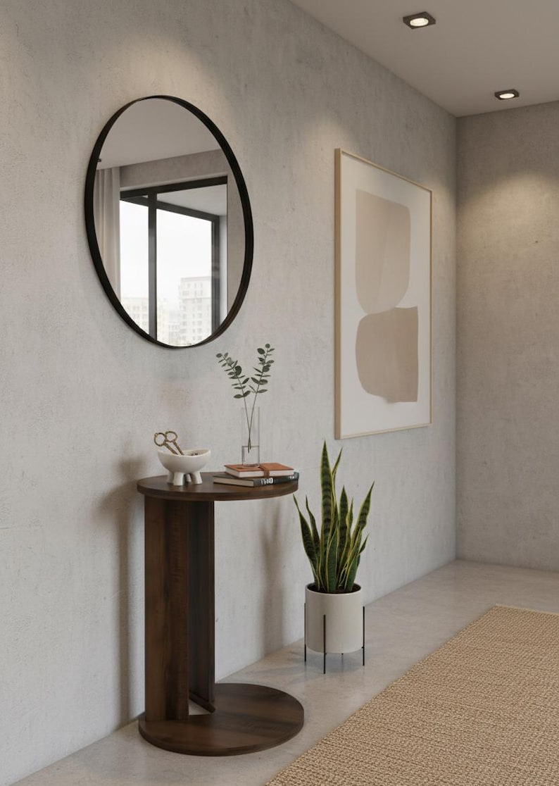 Op de afbeelding: Een moderne interieursc&egrave;ne met een donkerhouten bijzettafel, een ovale spiegel en een slangplant in een pot. De tafel bevat decoratieve items en boeken. Een abstract kunstwerk in een lijst hangt aan de muur.