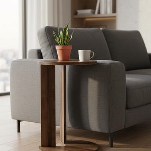 Op de afbeelding: Een bijzettafel van donker hout met een rond blad en een gebogen verticale steunvoet. Op de tafel staat een kleine groene sansevieria in een terracotta pot naast een effen witte keramische mok.
