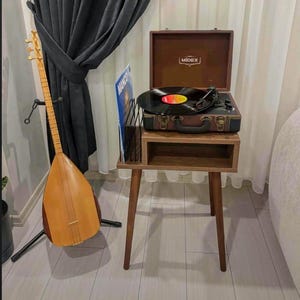 Puede incluir: Un tocadiscos de estilo vintage con un mueble y patas de madera marrón. El tocadiscos está reproduciendo un disco de vinilo. Un instrumento de cuerda de madera está en el fondo. El tocadiscos tiene un altavoz incorporado y un asa.