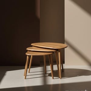 Op de afbeelding: Drie lichtbruine, ovale nesttafels met slanke, taps toelopende poten. De tafels zijn in aflopende volgorde gerangschikt. De tafels zijn gemaakt van hout en hebben een gladde afwerking. De achtergrond is neutraal. De kleinste tafel is ongeveer 40 cm hoog.