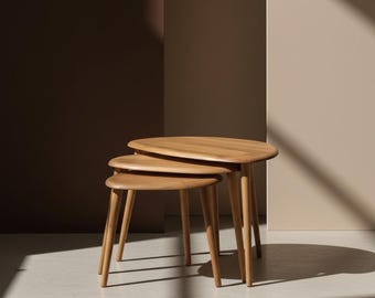 Puede incluir: Tres mesas nido ovaladas de color marrón claro con patas delgadas y cónicas. Las mesas están dispuestas en orden descendente. Las mesas son de madera y tienen un acabado liso. El fondo es neutro. La mesa más grande mide unos 60 cm de largo.