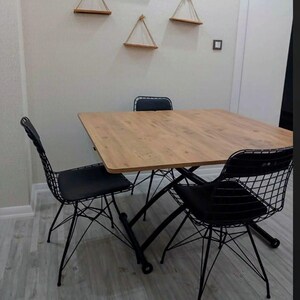 May include: A square wooden dining table with a black metal frame and four black wire mesh chairs. The table has a light wood grain finish. The chairs have black padded seats.