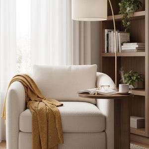 Op de afbeelding: Een gezellige woonkamer met een cr&egrave;mekleurige fauteuil en een mosterdgele deken. Een bijzettafel met een boek, bril en een kopje koffie. Een gouden booglamp en een houten boekenkast maken de setting compleet.