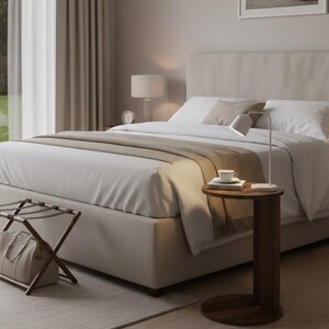Op de afbeelding: Een slaapkamerinterieur met een bed met wit beddengoed en een beige hoofdbord. Een kleine, ronde, donkere houten bijzettafel heeft een lamp, een kopje en boeken. Een bagagerek en een ingelijste landschapsfoto zijn ook zichtbaar.