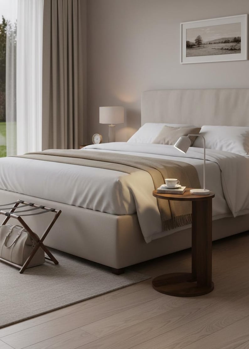 Op de afbeelding: Een slaapkamerinterieur met een bed met wit beddengoed en een beige hoofdbord. Een kleine, ronde, donkere houten bijzettafel heeft een lamp, een kopje en boeken. Een bagagerek en een ingelijste landschapsfoto zijn ook zichtbaar.