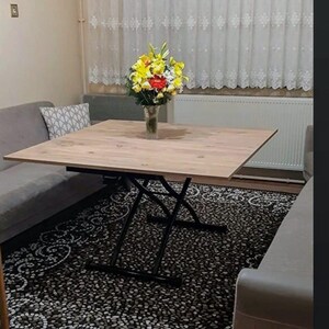 May include: A light wood square table with black metal legs that extend to raise the table height. The table is set on a black and white patterned rug in a living room setting.