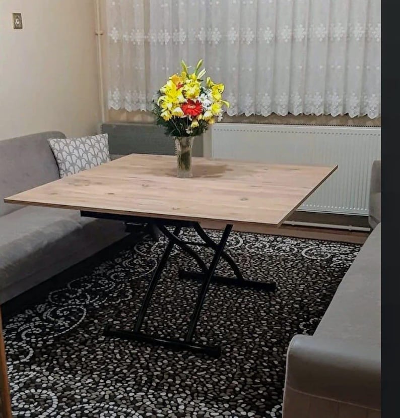 May include: A light wood square table with black metal legs that extend to raise the table height. The table is set on a black and white patterned rug in a living room setting.