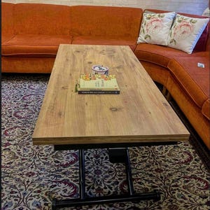 Peut inclure: Une table basse en bois à plateau relevable avec un cadre en métal noir et des roulettes. La table est relevée à une hauteur debout et a deux livres et une tasse en céramique blanche sur le dessus.
