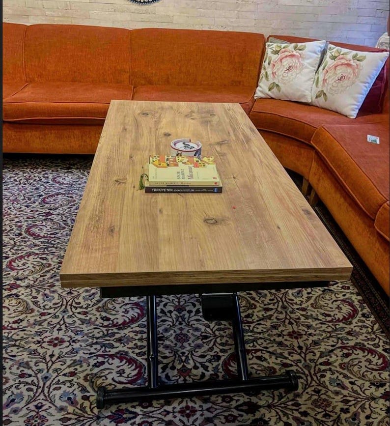 May include: A wooden lift-top coffee table with a black metal frame and wheels. The table is raised to a standing height and has two books and a white ceramic cup on top.