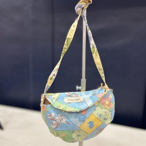May include: A patchwork shoulder bag with a flap. The bag is made of pastel-coloured squares with animal and floral patterns. The strap has a matching floral print. The text "Stoned Seagulls" is visible on the flap.