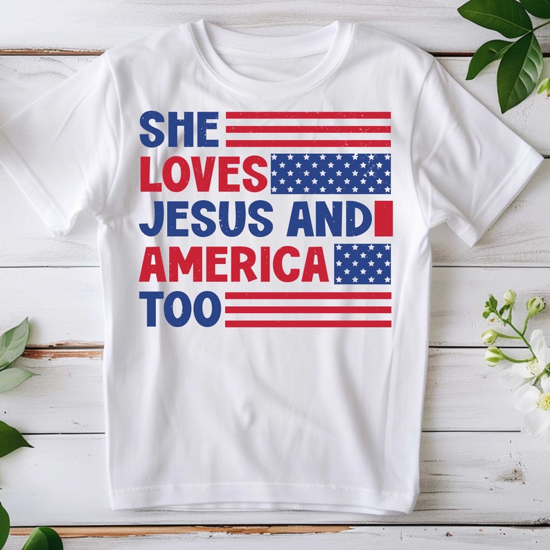 May include: White t-shirt with the text "SHE LOVES JESUS AND AMERICA TOO" in red and blue. The design incorporates American flag elements, with stripes and stars. The shirt is a casual wear item.