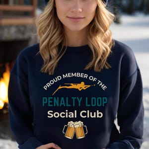 May include: Navy blue sweatshirt with the text "PROUD MEMBER OF THE PENALTY LOOP Social Club." The design includes a skier in yellow and two beer mugs. The sweatshirt is a comfortable, casual style.
