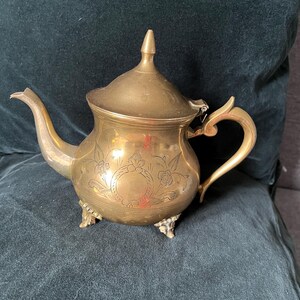 Pode incluir: Uma chaleira de latão com corpo arredondado, padrão floral gravado, tampa pontiaguda, pega curva e bico longo. A chaleira assenta sobre três pequenos pés decorativos e apresenta um acabamento metálico polido com uma pátina visível.