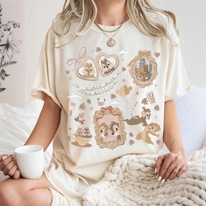 May include: A cream-coloured short-sleeve t-shirt featuring a brown graphic print of Chip and Dale characters. The design includes heart-shaped frames, a decorative mirror, and the text "DINED WITH MY BESTIE" surrounded by small icons like acorns.
