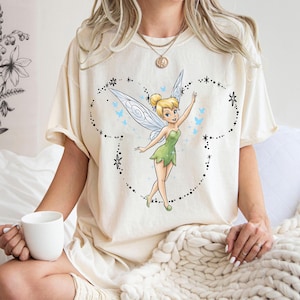 May include: A cream-coloured short-sleeve t-shirt featuring a graphic of Tinker Bell with translucent wings, surrounded by a dotted outline of Mickey Mouse ears, small blue butterflies, and star accents. The design is centred on the chest.