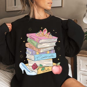 Op de afbeelding: Een zwart sweatshirt met ronde hals en een afbeelding van een stapel boeken met de namen Aurora, Erana, Snow White, Cinderella en Belle op de ruggen. Bovenop ligt een gouden tiara, met een glazen muiltje en een rode appel aan de onderkant.