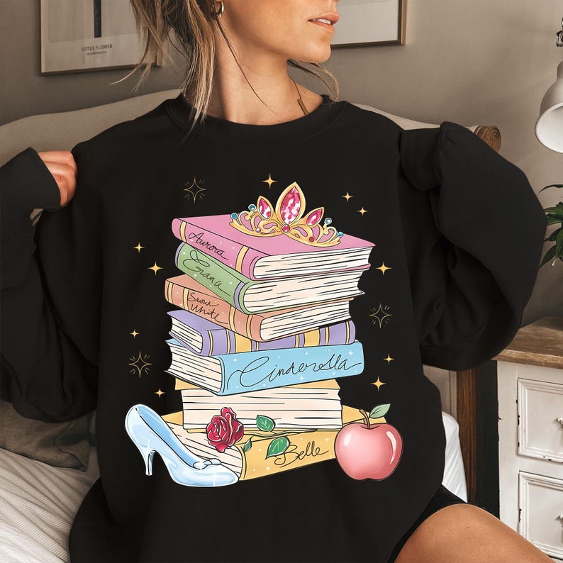 Op de afbeelding: Een zwart sweatshirt met ronde hals en een afbeelding van een stapel boeken met de namen Aurora, Erana, Snow White, Cinderella en Belle op de ruggen. Bovenop ligt een gouden tiara, met een glazen muiltje en een rode appel aan de onderkant.