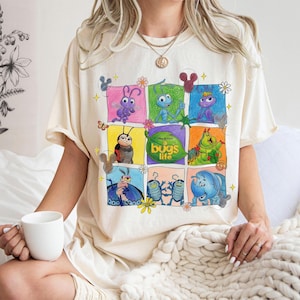Può includere: Una t-shirt a maniche corte color crema con una griglia di illustrazioni colorate dei personaggi del film A Bug's Life. La grafica include vari insetti in riquadri quadrati con il testo A Bug's Life centrato su una foglia verde.