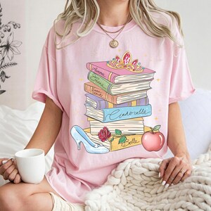 Op de afbeelding: Een lichtroze T-shirt met een afbeelding van een stapel boeken met de namen Aurora, Elana, Snow White, Cinderella en Belle op de ruggen. Bovenop ligt een gouden tiara, met een blauwe glazen muiltje en een rode appel aan de onderkant.
