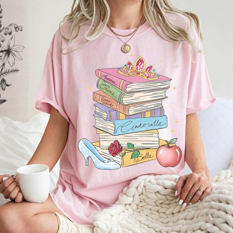 Op de afbeelding: Een lichtroze T-shirt met een afbeelding van een stapel boeken met de namen Aurora, Elana, Snow White, Cinderella en Belle op de ruggen. Bovenop ligt een gouden tiara, met een blauwe glazen muiltje en een rode appel aan de onderkant.