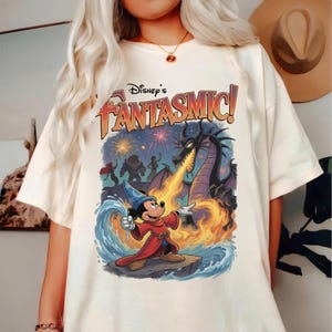 May include: A cream-colored t-shirt featuring a graphic print of Mickey Mouse as a sorcerer in a red robe and blue hat, casting a wave of water at a large black dragon breathing fire. The text Disney's Fantasmic! is printed above the scene.