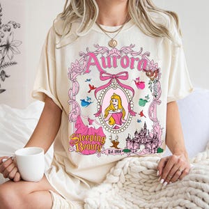 May include: A cream-colored short-sleeve t-shirt featuring a graphic print of Aurora from Sleeping Beauty. The design includes a pink ornate frame, the text Aurora and Sleeping Beauty Est. 1959, a castle, small birds, and fairies in pink, blue, and green.
