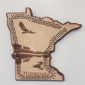 May include: Wooden cribbage board in the shape of Minnesota, featuring an eagle, a loon, and the word "Minnesota." The board has a natural wood finish with dark brown accents and numerous holes for pegs.