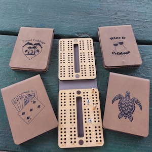 May include: Four tan leather-like folding travel cribbage boards with various black graphic designs including palm trees, wine glasses, playing cards, and a sea turtle, alongside one open board featuring a light wood pegging track.