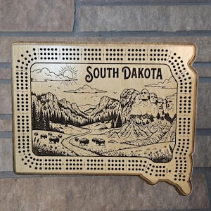 May include: A light-colored wooden cribbage board shaped like the state of South Dakota. The board features a laser-engraved landscape illustration of Mount Rushmore, bison, and a sun, with the text South Dakota printed at the top center.