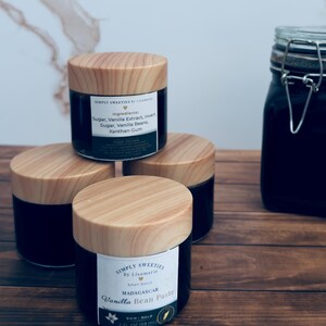 May include: Four small jars of Madagascar Vanilla Bean Paste with wooden lids are stacked on a wooden surface. Each jar has a label with the text "Simply Sweeties" and ingredient information. A larger jar is visible in the background.