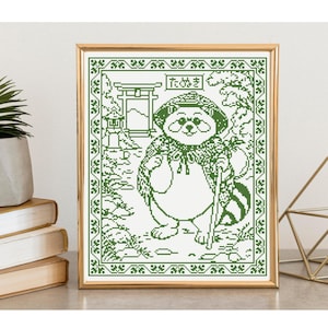 May include: A framed cross-stitch pattern in dark green thread on a white background, depicting a tanuki character wearing a hat and cape, holding a staff, with a torii gate and Japanese text that reads "たぬき" inside a decorative border.