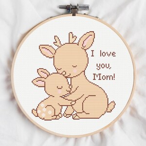 May include: A cross-stitch hoop featuring a mother deer and fawn in shades of tan and pink, with the text "I love you, Mom!" in brown. The design is set against a white background.