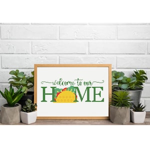 May include: A framed cross-stitch design with the words "welcome to our HOME" in green. The "O" in HOME is replaced with a yellow taco with red and green accents. The frame is a light brown color. The design is displayed in front of a white brick wall.