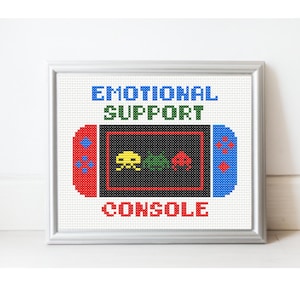 May include: Cross-stitch artwork in a silver frame. The design shows a pixelated video game console with the words "EMOTIONAL SUPPORT CONSOLE". The console has red and blue sides with a black screen displaying yellow, green, and red pixelated characters.