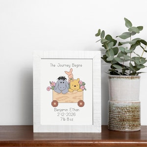 Könnte beinhalten: Ein gerahmtes Kreuzstichbild mit Winnie-the-Pooh-Figuren in einem Holzwagen mit Blumen. Der Text "The Journey Begins" steht über dem Wagen. Darunter steht "Benjamin Ethan 2-12-2026 3,4 kg". Der Rahmen ist weiß.
